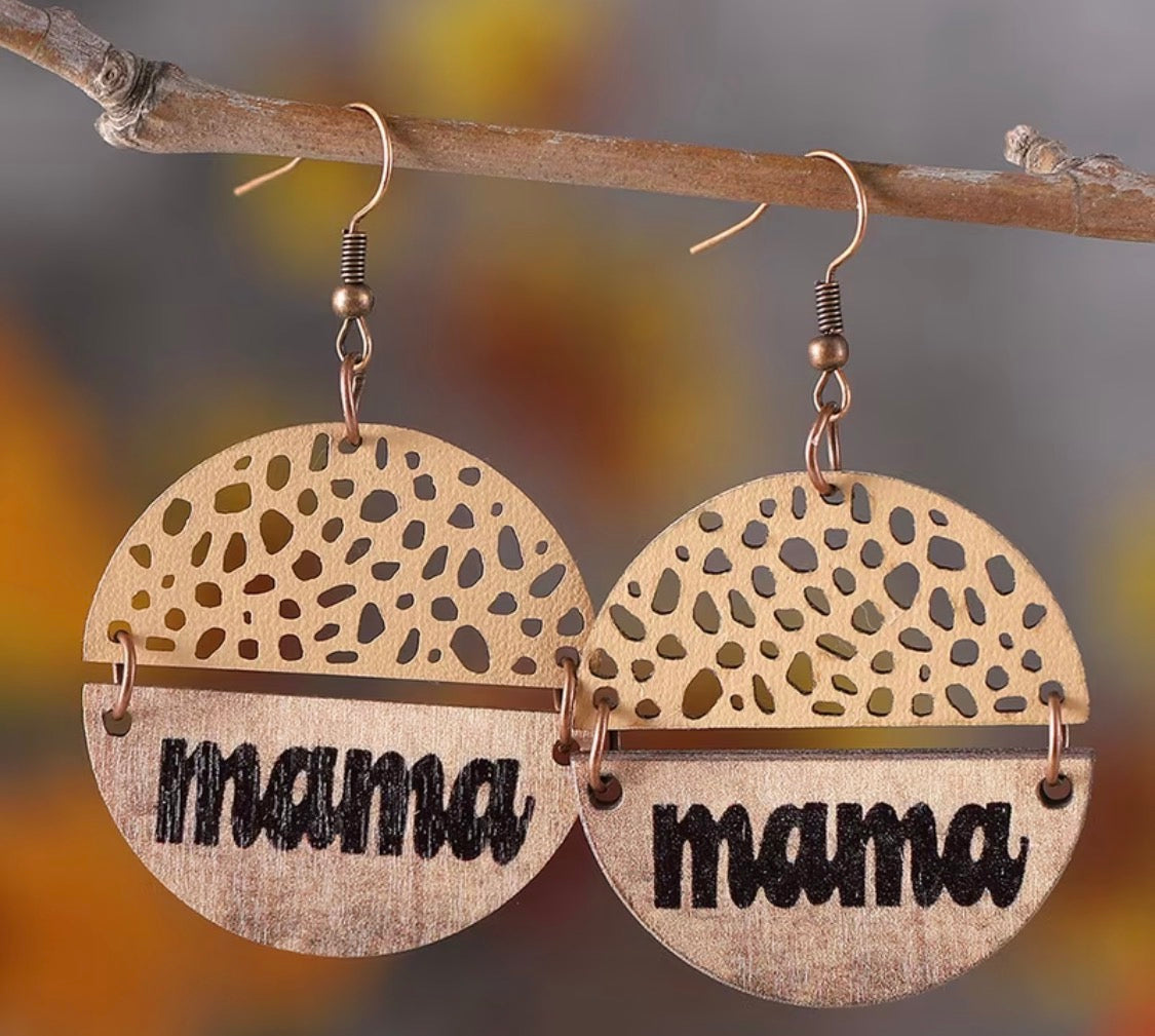 A pair of round wooden dangle earrings hanging from a small branch. Each earring has a laser-cut leopard-like spot pattern on the top half and the word "mama" in black script on the bottom half.